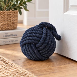 May include: A dark navy blue doorstop crafted from thick, tightly coiled rope tied into a spherical monkey's fist knot. The heavy, textured nautical-style knot sits on a wooden floor, positioned to hold a white door open with a single loose end.