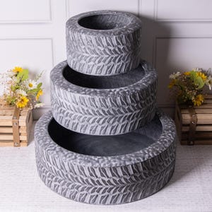 May include: Three grey, textured tyre-shaped planters stacked. The planters have a detailed tread pattern and are of varying sizes. The background includes a white wall and wooden crates with flowers.