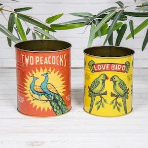 May include: Two cylindrical metal tins with vintage-style graphics. The larger orange tin features two peacocks and the text "TWO PEACOCKS". The smaller yellow tin displays two green parrots on a branch with the text "LOVE BIRD". Both tins have visible weight text.