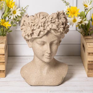 Può includere: Un vaso color pietra beige a forma di testa umana, caratterizzato da una corona scolpita di vari fiori sulla fronte. Il busto presenta una consistenza liscia e punteggiata e un'espressione facciale serena con occhi chiusi e un lieve sorriso.
