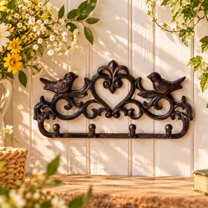 May include: A dark brown, cast iron wall-mounted hook rack featuring a decorative scrollwork design with a central heart shape and two birds perched on the ends. The rack includes four rounded hooks along the bottom edge for hanging items.