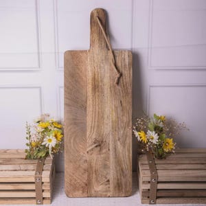 May include: A rectangular wooden chopping board with a handle and a rope loop for hanging. The board is made of light brown wood with visible grain and knots. The handle is attached with a rope. The board is displayed with flowers.