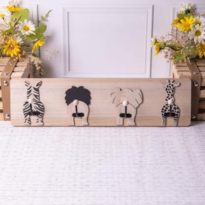 May include: Wooden wall-mounted coat rack featuring four animal designs: a zebra, a lion, an elephant, and a giraffe. Each animal has a hook for hanging items. The rack is made of light-colored wood and is decorated with black and white details.