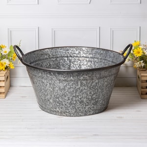 May include: A large, oval galvanised metal tub with black handles. The tub has a textured, mottled appearance and a dark rim. The background features a white wall and wooden crates with yellow and white flowers.