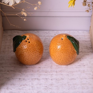 May include: A pair of ceramic salt and pepper shakers shaped like oranges. The shakers are orange with a textured surface and green leaf accents. Each shaker has holes on top for dispensing. A decorative kitchen item.