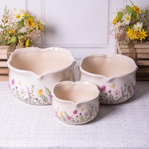 May include: A trio of off-white ceramic plant pots with a heart-shaped rim. Each pot is adorned with a floral design in purple, yellow, and green. The pots are different sizes, ideal for various plants. Measurements are in centimeters.