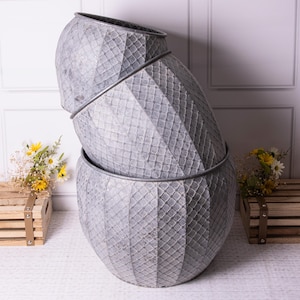 May include: Three grey metal planters stacked together, each with a diamond pattern. The planters have a rounded shape and a silver rim. They are arranged in a leaning tower style, creating a unique display.