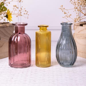 Könnte beinhalten: Drei Glasvasen mit gerippten Texturen. Die Vase links ist lila, die mittlere Vase ist gelb und die Vase rechts ist grau.