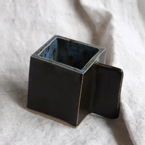 May include: A black ceramic mug with a square shape and a handle on the side. The mug has a blue and black glaze.