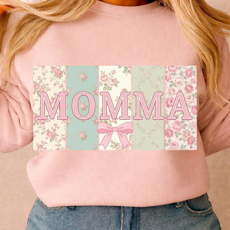 K&ouml;nnte beinhalten: Hellrosa Sweatshirt mit dem Wort "MOMMA" in rosa Buchstaben. Die Buchstaben sind vor einem floralen Muster aus rosa Rosen und gr&uuml;nen Bl&auml;ttern platziert. Unter den Buchstaben befindet sich eine rosa Schleife.