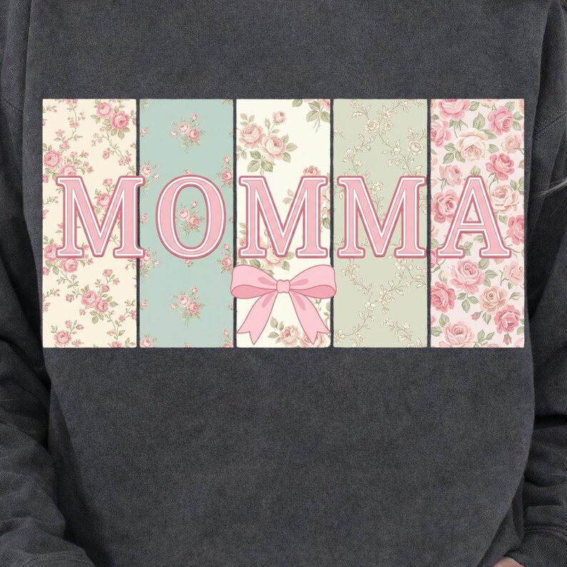 K&ouml;nnte beinhalten: Dunkelgraues Sweatshirt mit dem Wort "MOMMA" in rosa Buchstaben mit floralem Muster. Die Buchstaben sind vor einem Hintergrund aus pastellfarbenen Blumenmustern platziert, mit einer rosa Schleife unter dem "O".