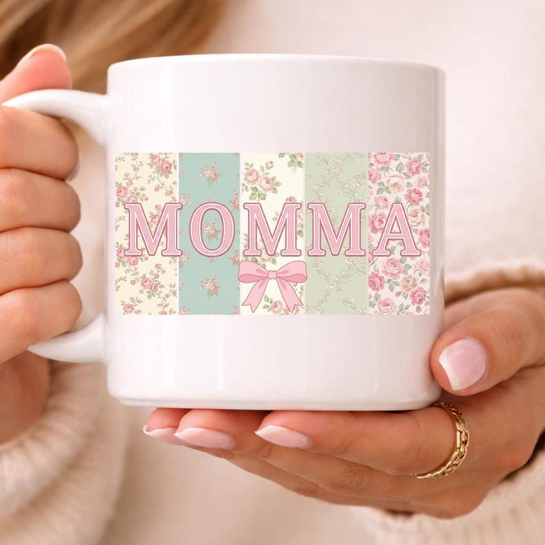 K&ouml;nnte beinhalten: Wei&szlig;e Keramiktasse mit dem Wort "MOMMA" in rosa Blockbuchstaben. Die Buchstaben sind vor einem floralen Patchwork-Design in Pastellfarben, darunter Rosa, Gr&uuml;n und Creme, platziert. Unter dem Text "MOMMA" befindet sich eine rosa Schleife.