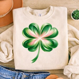 May include: Cream-colored sweatshirt featuring a large, stylized four-leaf clover design. The clover has green and pink hues, with detailed shading. The sweatshirt is made of a soft material and is suitable for casual wear.