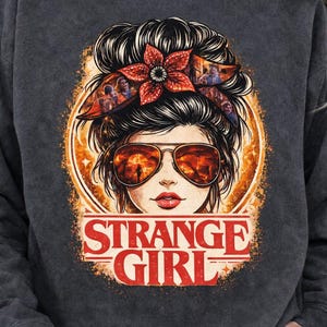 May include: Dark gray sweatshirt featuring a graphic of a woman with sunglasses and a floral headband. The text "STRANGE GIRL" is displayed in red. The design has an orange and brown color scheme.
