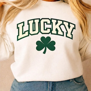 May include: A white sweatshirt with the word "LUCKY" in green block letters with a cream outline. Below the word "LUCKY" is a green shamrock. The sweatshirt is a casual style.