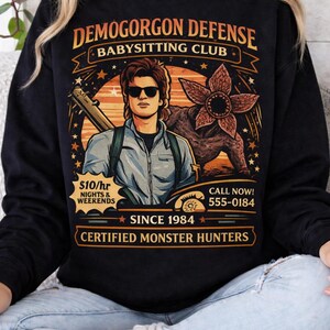 May include: Black sweatshirt with a graphic featuring a man with sunglasses, a Demogorgon, and text that reads "DEMOGORGON DEFENSE BABYSITTING CLUB." Additional text includes "$10/hr NIGHTS & WEEKENDS," "SINCE 1984," and "CERTIFIED MONSTER HUNTERS."