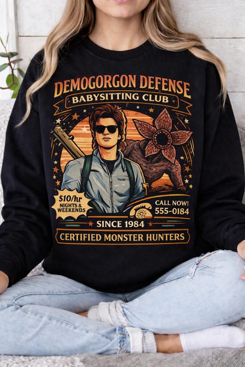 May include: Black sweatshirt with a graphic featuring a man with sunglasses, a Demogorgon, and text that reads "DEMOGORGON DEFENSE BABYSITTING CLUB." Additional text includes "$10/hr NIGHTS & WEEKENDS," "SINCE 1984," and "CERTIFIED MONSTER HUNTERS."
