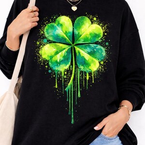Peut inclure: Sweat-shirt noir orné d'un motif de trèfle à quatre feuilles de style aquarelle. Le trèfle est vert et jaune, avec des effets de peinture dégoulinante. Le motif est centré sur le devant du vêtement.