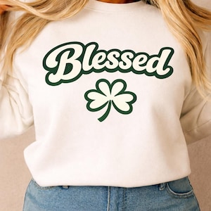 May include: White sweatshirt with the word "Blessed" in a green, retro font, and a green and white shamrock graphic. The sweatshirt is a casual style, suitable for St. Patrick's Day or everyday wear.