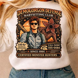 May include: White sweatshirt with a graphic featuring a man with sunglasses, a Demogorgon, and the text "DEMOGORGON DEFENSE BABYSITTING CLUB." Includes the text "$10/hr NIGHTS & WEEKENDS" and "CERTIFIED MONSTER HUNTERS."