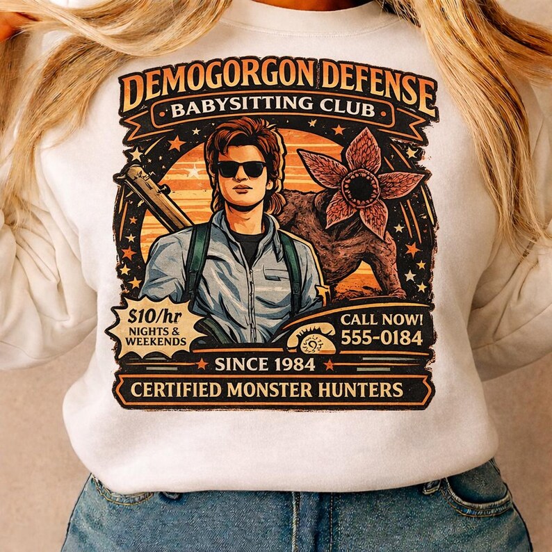 May include: White sweatshirt with a graphic featuring a man with sunglasses, a Demogorgon, and the text "DEMOGORGON DEFENSE BABYSITTING CLUB." Includes the text "$10/hr NIGHTS & WEEKENDS" and "CERTIFIED MONSTER HUNTERS."