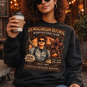 May include: Black sweatshirt with the text "DEMOGORGON DEFENSE BABYSITTING CLUB" and a graphic of a man and a monster. The sweatshirt also has the text "$10/hr Nights & Weekends" and "Certified Monster Hunters".