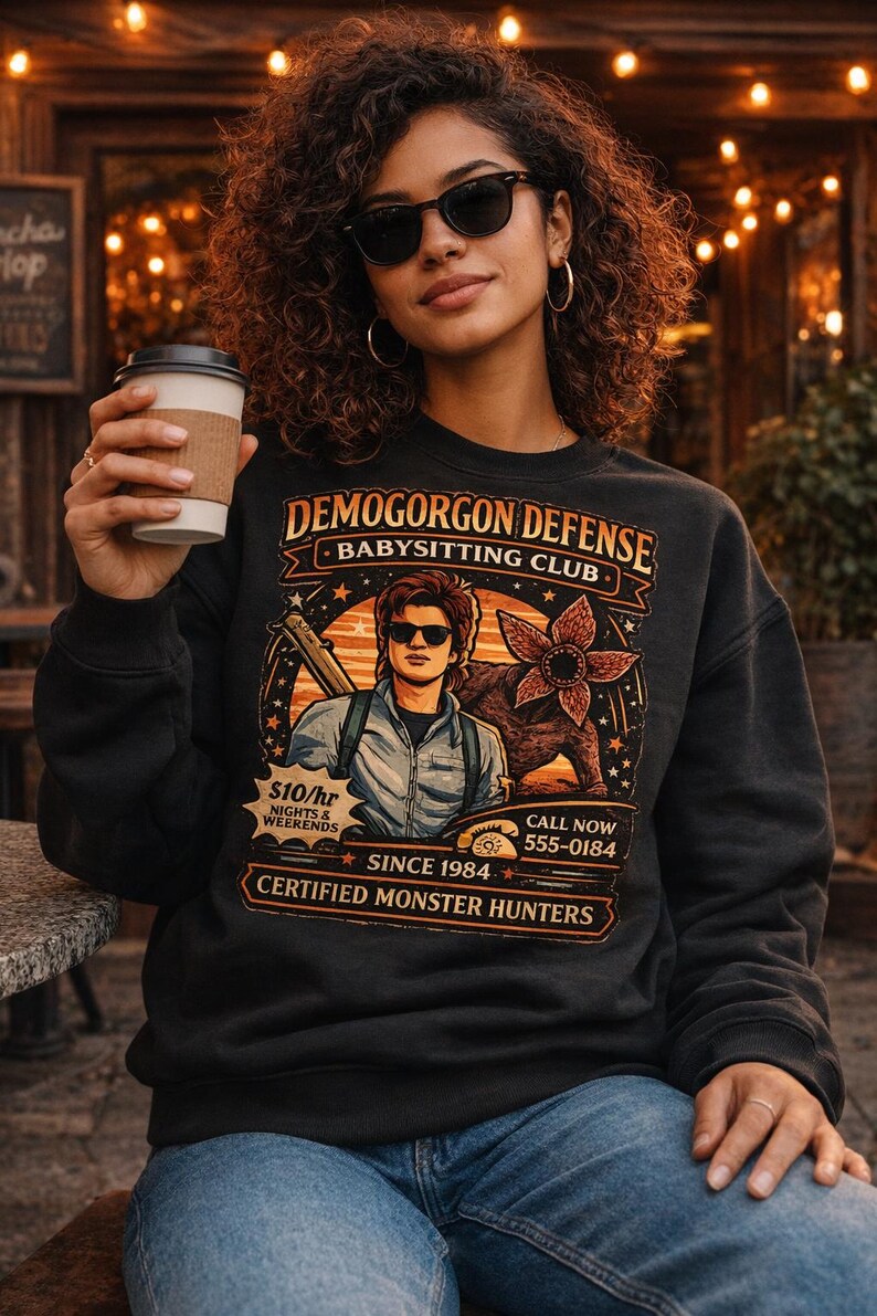 May include: Black sweatshirt with the text "DEMOGORGON DEFENSE BABYSITTING CLUB" and a graphic of a man and a monster. The sweatshirt also has the text "$10/hr Nights & Weekends" and "Certified Monster Hunters".
