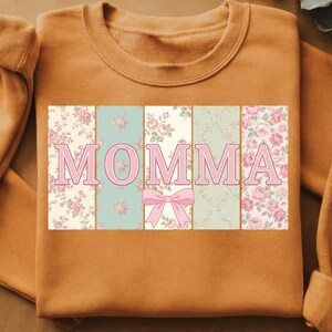 K&ouml;nnte beinhalten: Ein orangefarbenes Sweatshirt mit dem Wort "MOMMA" in Rosa. Die Buchstaben sind vor einem floralen Patchwork-Design mit rosa Rosen und einer rosa Schleife platziert.