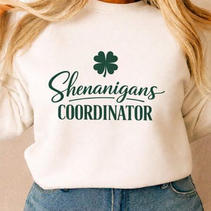 Peut inclure: Sweat-shirt blanc avec l'inscription "Shenanigans COORDINATOR" en vert foncé, orné d'un trèfle vert. Vêtement de style décontracté.