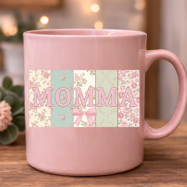 K&ouml;nnte beinhalten: Eine rosa Keramiktasse mit dem Wort "MOMMA" in einem floralen Design. Die Buchstaben sind vor einem Hintergrund aus pastellfarbenen Blumenmustern mit einer rosa Schleife angeordnet. Die Tasse hat einen gebogenen Griff.