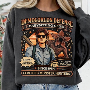 May include: Dark gray sweatshirt with a graphic featuring the text "DEMOGORGON DEFENSE BABYSITTING CLUB." The design includes an illustration of a person with sunglasses, a Demogorgon, and the text "CERTIFIED MONSTER HUNTERS."