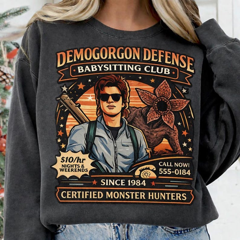 May include: Dark gray sweatshirt with a graphic featuring the text "DEMOGORGON DEFENSE BABYSITTING CLUB." The design includes an illustration of a person with sunglasses, a Demogorgon, and the text "CERTIFIED MONSTER HUNTERS."