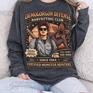 May include: Dark gray long-sleeve shirt with a graphic featuring the text "DEMOGORGON DEFENSE BABYSITTING CLUB" and an illustration of a man with sunglasses and a monster. The shirt also includes the text "SINCE 1984" and "CERTIFIED MONSTER HUNTERS".