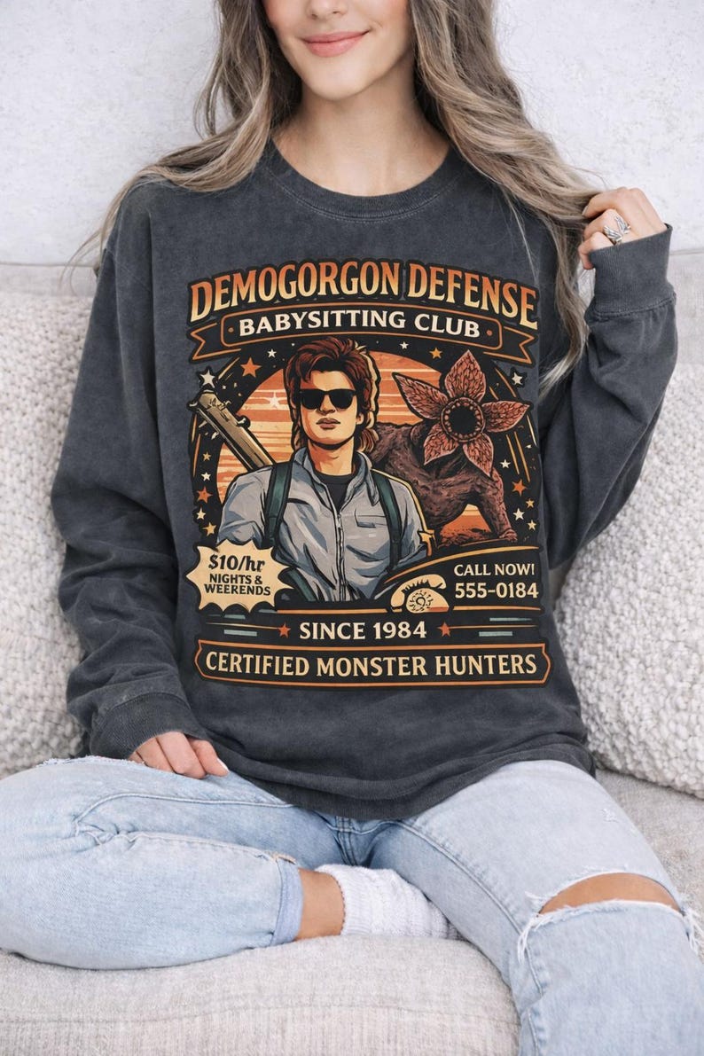 May include: Dark gray long-sleeve shirt with a graphic featuring the text "DEMOGORGON DEFENSE BABYSITTING CLUB" and an illustration of a man with sunglasses and a monster. The shirt also includes the text "SINCE 1984" and "CERTIFIED MONSTER HUNTERS".