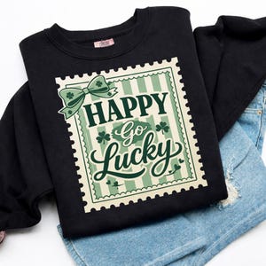 Puede incluir: Sudadera negra con un gran gráfico con las palabras "HAPPY Go Lucky" en un diseño tipo sello. El diseño incluye un lazo verde, tréboles y un fondo a rayas. La sudadera está doblada sobre unos pantalones cortos vaqueros azules.