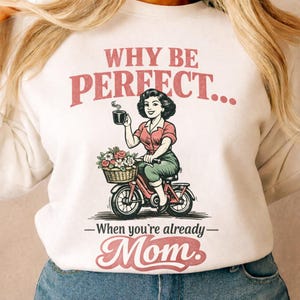 Könnte beinhalten: Weißes Sweatshirt mit einer Grafik einer Frau auf einem roten Fahrrad, die eine Kaffeetasse und einen Blumenkorb hält. Der Text lautet: „WARUM PERFEKT SEIN... Wenn du schon Mama bist.“