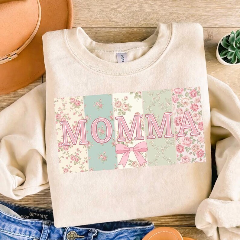 K&ouml;nnte beinhalten: Cremefarbenes Sweatshirt mit dem Wort "MOMMA" in rosa Buchstaben. Die Buchstaben sind vor einem Patchwork-Design aus floralen und pastellfarbenen Bl&ouml;cken angeordnet, mit einer rosa Schleife darunter.