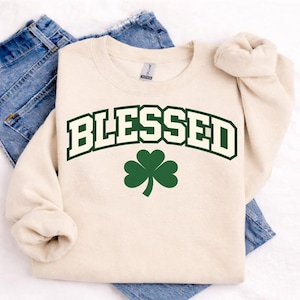 May include: A cream-colored sweatshirt with the word "BLESSED" in a green, collegiate-style font. Below the word is a green shamrock. The sweatshirt is folded on top of a pair of blue jeans.