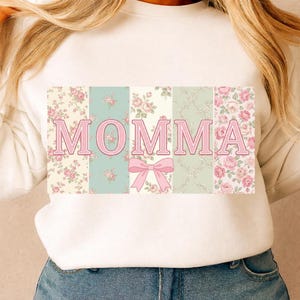 K&ouml;nnte beinhalten: Wei&szlig;es Sweatshirt mit dem Wort "MOMMA" in rosa Buchstaben. Die Buchstaben sind vor einem Patchwork aus Blumenmustern in Rosa, Gr&uuml;n und Creme platziert. Unter den Buchstaben befindet sich eine rosa Schleife.
