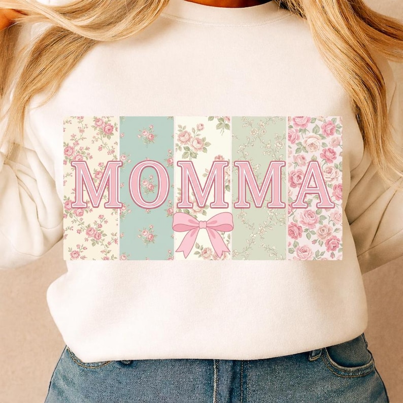K&ouml;nnte beinhalten: Wei&szlig;es Sweatshirt mit dem Wort "MOMMA" in rosa Buchstaben. Die Buchstaben sind vor einem Patchwork aus Blumenmustern in Rosa, Gr&uuml;n und Creme platziert. Unter den Buchstaben befindet sich eine rosa Schleife.