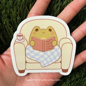 May include: A sticker featuring a cartoon frog wearing a blanket and reading a book titled "Cozy Stories" while sitting in a yellow armchair. The frog is green with a yellow belly and has a cup of tea on a small table next to the chair.