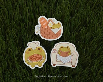 May include: Three frog-themed stickers. One sticker shows a frog wearing a chef's hat and holding a pie. Another sticker shows a frog sitting in a chair reading a book. The third sticker shows a frog sitting in a basket with bread and flowers.