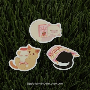 May include: Three sticker designs featuring cute cats with different Japanese snacks. One cat is holding a strawberry milk carton, another is holding a box of Pocky sticks, and the third is wearing a bag of potato chips.