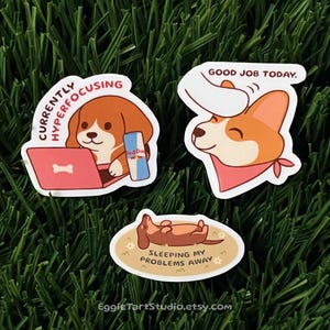 May include: Three cartoon dog stickers on a green background. One sticker shows a beagle with a laptop and a Red Dog energy drink, with the text "Currently Hyperfocusing." Another shows a corgi with the text "Good Job Today." The third shows a dachshund with the text "Sleeping My Problems Away."