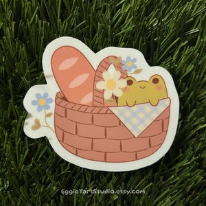 May include: A sticker of a pink picnic basket with a green frog peeking out, holding a blue and white checkered blanket. The basket is filled with a loaf of bread and flowers.