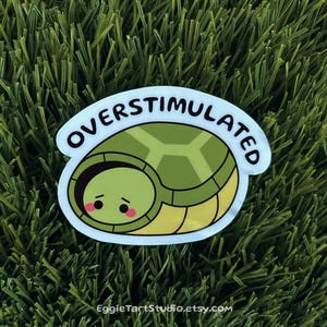 May include: A white sticker with a cartoon turtle with its head tucked inside its shell. The turtle is green and yellow and has a cute face. The sticker says "Overstimulated" in black text.