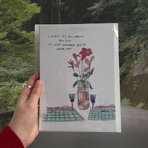 Peut inclure: Une carte blanche avec une illustration dessinée à la main d'un vase avec des fleurs rouges et deux verres de vin sur une nappe à carreaux. La carte porte le texte "Je suppose que tout est une question d'essayer d'aimer quelqu'un que l'on n'a jamais rencontré."