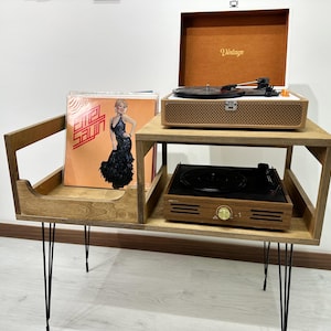 Puede incluir: Un soporte de madera para tocadiscos con un estante incorporado para guardar discos. El soporte tiene patas de horquilla negras y presenta un tocadiscos de estilo vintage en el estante superior. El tocadiscos tiene una caja de madera marrón con un plato giratorio negro y una perilla de control dorada.