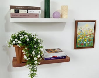 Può includere: Esposizione di articoli per la casa su mensole bianche. Include libri, candele, un piccolo dipinto di fiori e una pianta in vaso con fiori bianchi. Le mensole sono montate su un muro bianco.