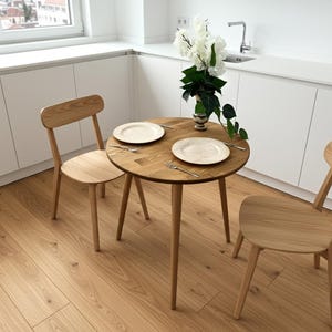 Puede incluir: Una pequeña mesa de comedor redonda de madera con dos sillas a juego. La mesa está puesta con dos platos, cubiertos y un jarrón con flores blancas. Las sillas y la mesa son de madera clara. La mesa tiene unos 80 cm de diámetro.